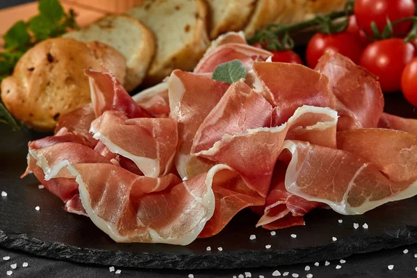 Krájený jamon, cherry rajčata, bageta, bylinky a koření na černé kamenné břidlicové desce na tmavě šedém pozadí. Záběr zblízka. Boční pohled. — Stock fotografie