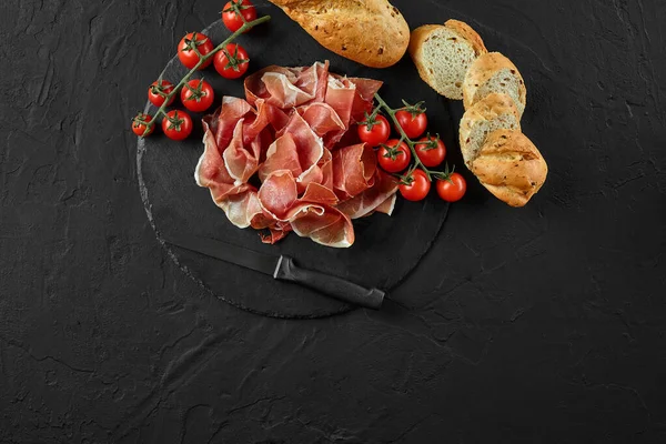 Jamon fatiado, tomates cereja, baguete fatiado e uma faca na placa de ardósia de pedra preta contra um fundo cinza escuro. Close-up. Vista superior. Copiar espaço. — Fotografia de Stock