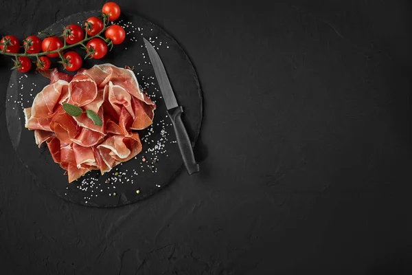 Plasterek jamonu, pomidory wiśniowe, zioła, przyprawy i nóż na tablicy z czarnego kamienia na ciemnoszarym tle. Strzał z bliska. Widok z góry. — Zdjęcie stockowe