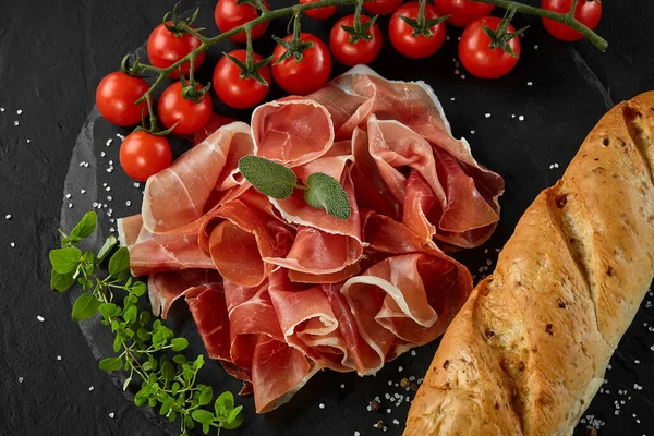 Krájený jamon, cherry rajčata, bageta, bylinky a koření na černé kamenné břidlicové desce na tmavě šedém pozadí. Záběr zblízka. Horní pohled. — Stock fotografie