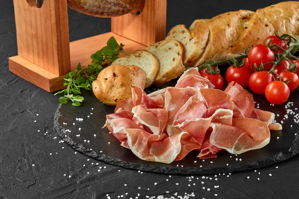 Jamon carved from pig leg on a jamonera stand, cherry tomatoes, baguette, herbs, spices on black slate board. Dark background. Close-up. Side view. — Stock Image Jamon carved from pig leg on a jamonera stand, cherry tomatoes, baguette, herbs, spices on black slate board. Dark background. Close-up. Side view. Stock Photo
