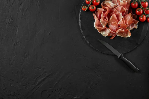 Sliced jamon, cherry tomatoes and a knife on black stone slate board against a dark grey background. Close-up shot. Top view. Copy space. — Stock Image Sliced jamon, cherry tomatoes and a knife on black stone slate board against a dark grey background. Close-up shot. Top view. Copy space. Stock Photo