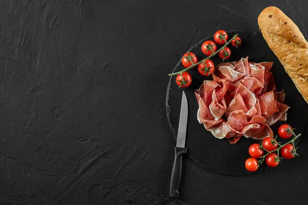 Sliced jamon, cherry tomatoes, baguette and a knife on black stone slate board against a dark grey background. Close-up shot. Top view. Copy space. — Stock Image Sliced jamon, cherry tomatoes, baguette and a knife on black stone slate board against a dark grey background. Close-up shot. Top view. Copy space. Stock Photo