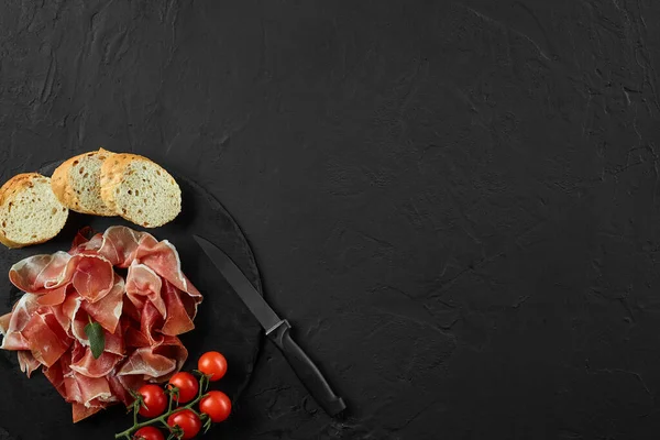 Sliced jamon, cherry tomatoes, baguette, herbs and knife on black stone slate board against dark grey background. Close-up. Top view. Copy space. — Stock Image Sliced jamon, cherry tomatoes, baguette, herbs and knife on black stone slate board against dark grey background. Close-up. Top view. Copy space. Stock Picture