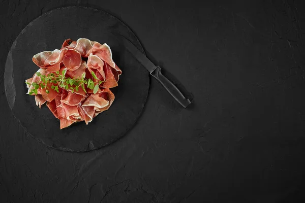 Sliced jamon with herbs and a knife are on black stone slate board against a dark grey background. Close-up shot. Top view. Copy space. — Stock Image Sliced jamon with herbs and a knife are on black stone slate board against a dark grey background. Close-up shot. Top view. Copy space. Stock Image