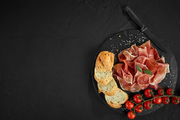 Sliced jamon, cherry tomatoes, baguette, herbs, spices and knife on black stone slate board against a dark grey background. Close-up shot. Top view. — Stock Image Sliced jamon, cherry tomatoes, baguette, herbs, spices and knife on black stone slate board against a dark grey background. Close-up shot. Top view. Royalty Free Stock Images