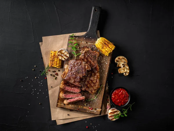 Smažené steaky s bylinkami a kořením na dřevěné desce, pergamen, grilovaný česnek, kukuřice, omáčka v tmavé míse na černém pozadí. Detailní záběr. Horní pohled. — Stock fotografie