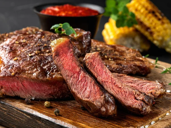 Geschnitten, gebraten, gewürzte Steaks mit Kräutern auf Holzbrett, gegrillter Mais, rote Soße in kleiner dunkler Schüssel auf schwarzem Hintergrund. Nahaufnahme. Seitenansicht. — Stockfoto