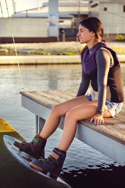 Femminile in dolcevita grigia, giubbotto sportivo speciale e pantaloncini in denim è in posa con un wakebord sulle gambe seduto su un molo di una zona costiera. Primo piano. — Foto Stock