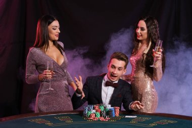 Kumarhanede poker oynayan zarif bir adam ve yanında iki kız var. Üstünde bir yığın fiş ve kartla masada oturmak. Siyah, duman arkaplanı.