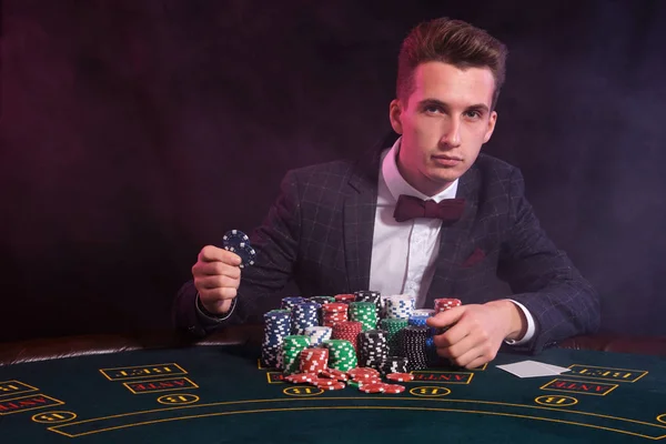 L'uomo elegante sta giocando a poker al casinò. È seduto al tavolo con pile di patatine colorate e carte su di esso. Nero, sfondo fumo . — Foto Stock