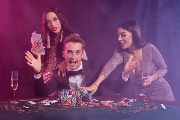 Uomo elegante che gioca a poker al casinò accompagnato da due ragazze. Seduto al tavolo con pile di patatine e carte sopra. Nero, sfondo fumo . — Foto Stock