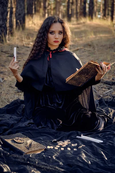 Bruxa em vestido preto com capa e capuz. Possuindo em floresta de pinheiro. Sentado em livros brancos escuros, velas e runas sobre ele. Comprimento total. — Fotografia de Stock