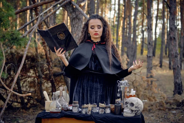 Häxa i svart klänning med cape och huva. Hålla bok, uttala förtrollning, poserar i tallskog vid bordet med tillbehör för trolldom. Närbild. — Stockfoto