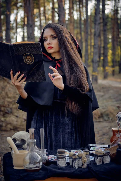 Bruxa em vestido preto com capa e capuz. Segurando livro, pronunciando feitiço, posando em pinhal à mesa com acessórios para bruxaria. Close-up. — Fotografia de Stock