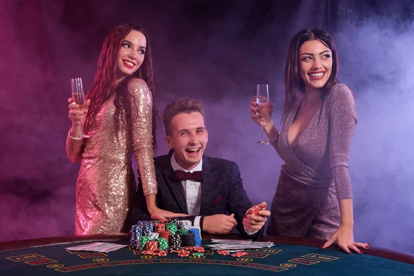 Eleganter Mann, der in Begleitung zweier Mädchen im Casino pokert. Sitzend am Tisch mit Stapeln von Chips und Karten darauf. Schwarz, rauchiger Hintergrund. — Stockfoto