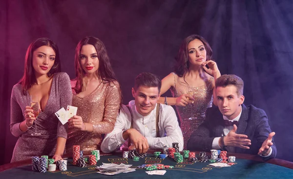 Des amis jouant au poker au casino. Ils célèbrent leur victoire, boivent du champagne et posent à table avec des piles de jetons, d'argent et de cartes dessus . — Photo