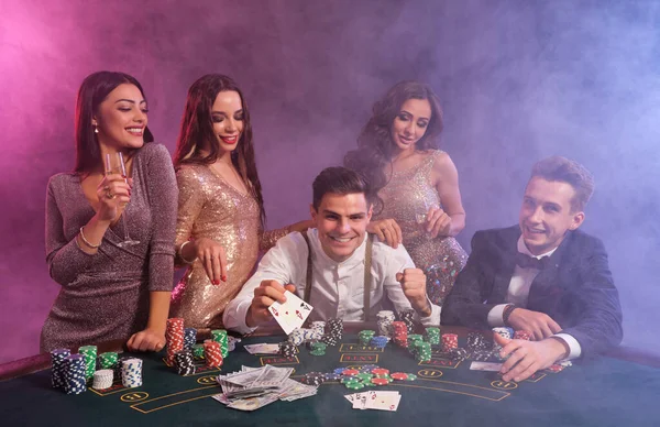 Przyjaciele grający w pokera w kasynie. Świętują swoje zwycięstwo, piją szampana i pozują przy stole ze stosami żetonów, pieniędzy i kart.. — Zdjęcie stockowe