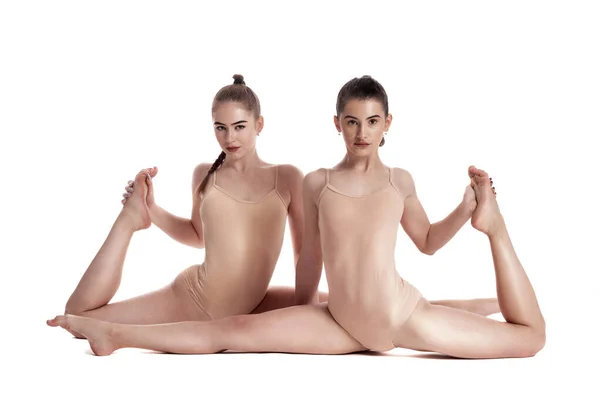 Δύο ευέλικτα κορίτσια γυμναστές με κοτσίδες, σε μπεζ λεοπαρδάλεις εκτελούν σχισμές ενώ θέτουν απομονωμένες σε λευκό φόντο. Κοντινό πλάνο. — Φωτογραφία Αρχείου