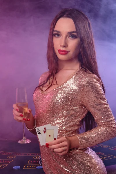 Mujer en vestido dorado mostrando cartas, sosteniendo champán mientras posando en la mesa de juego en el casino. Negro, fondo de humo. Juego, póquer. Primer plano . —  Fotos de Stock