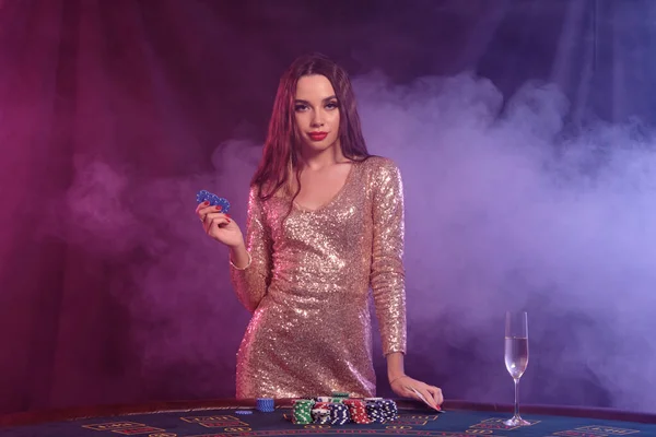 A lány arany ruhában zsetonokat mutat, miközben asztalnál pózol a kaszinóban. Fekete, füstös háttér, színes háttérvilágítás, pezsgő. Póker. Közelkép. — Stock Fotó