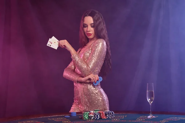 Frau in goldenem Kleid zeigt Chips und Karten, während sie am Spieltisch im Casino posiert. Schwarzer, rauchiger Hintergrund. Glücksspiel, Poker, Champagner. Nahaufnahme. — Stockfoto
