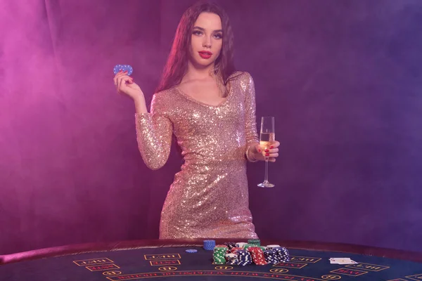 Žena ve zlatých šatech ukazuje chipsy, drží šampaňské, zatímco pózuje u hracího stolu v kasinu. Černá, kouřové pozadí. Hazard, poker. Detailní záběr. — Stock fotografie