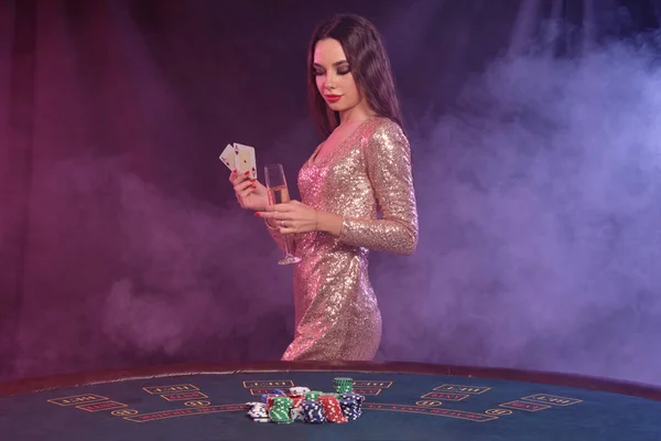 Frau in goldenem Kleid zeigt Karten, hält Champagner in der Hand, während sie am Spieltisch im Casino posiert. Schwarzer, rauchiger Hintergrund. Glücksspiel, Poker. Nahaufnahme. — Stockfoto