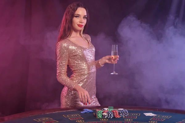 Woman in golden dress holding chip and champagne while posing at playing table in casino. Black, smoke background. Gambling, poker. Close-up. Royalty Free Stock Images