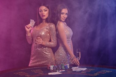 Kumarhanede oyun masasında poz veren iki muhteşem kadın. Siyah, renkli arka ışıkları olan dumanlı arka plan. Kumar, poker. Yakın plan..