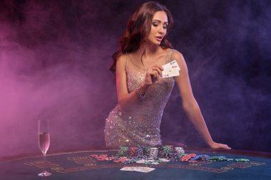 Poker oynayan bir kız, kumarhane. İskambil kağıtları göstermek, masada para ve çiplerle poz vermek. Siyah, dumanlı arka plan, renkli arka ışıklar. Yakın plan..
