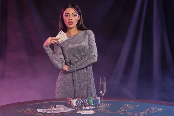 Žena v šedých šatech ukazuje karty, pózuje u hracího stolu v kasinu. Černé, kouřové pozadí s barevnými podsvíceními. Hazard, poker. Detailní záběr. — Stock fotografie