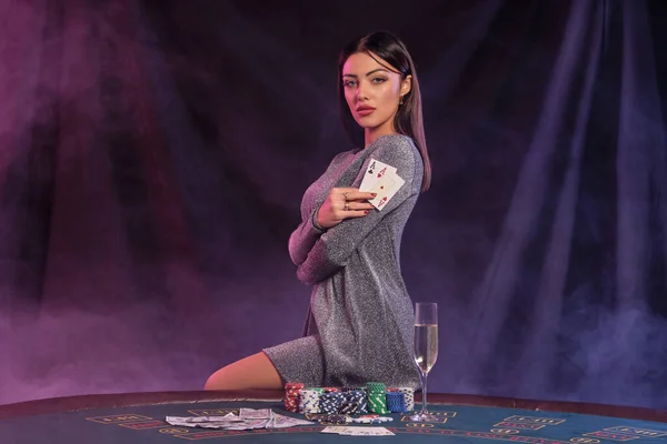 Žena v šedých šatech ukazuje karty, sedí na hracím stole v kasinu. Černé, kouřové pozadí s barevnými podsvíceními. Hazard, poker. Detailní záběr. — Stock fotografie