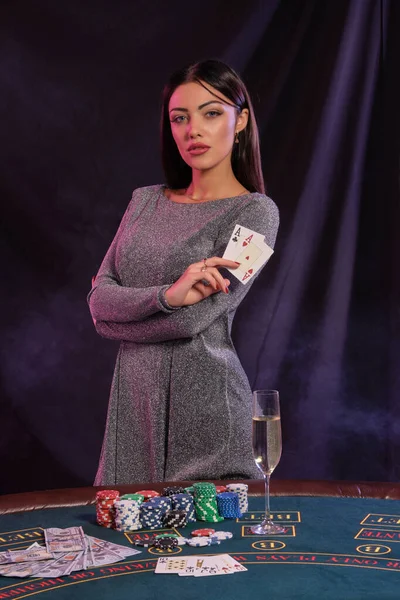 Žena v šedých šatech ukazuje karty, pózuje u hracího stolu v kasinu. Černé, kouřové pozadí s barevnými podsvíceními. Hazard, poker. Detailní záběr. — Stock fotografie