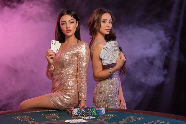 Две женщины показывают карты и деньги, позируют за игровым столом в казино. Черный, дымный фон с цветными подсветками. Азартные игры, покер. Закрытие. — стоковое фото