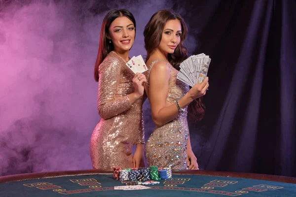 Δύο γυναίκες να δείχνουν χαρτιά και χρήματα, να ποζάρουν στο καζίνο. Μαύρο, φόντο καπνού με πολύχρωμα backlights. Τζόγος, πόκερ. Κοντινό πλάνο. — Φωτογραφία Αρχείου