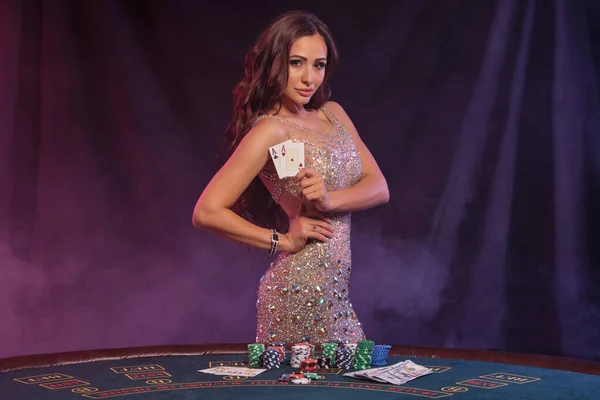 Menina jogando poker, cassino. Mostrando cartões, posando à mesa com pilhas de fichas e dinheiro. Preto, fundo de fumaça, backlights coloridos. Close-up. — Fotografia de Stock
