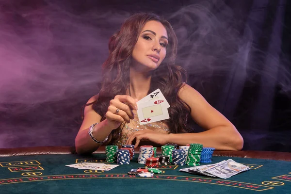 Flicka spelar poker, kasino. Visar kort, sitter vid bordet med högar av marker och pengar. Svart, rök bakgrund, färgglada bakljus. Närbild. — Stockfoto
