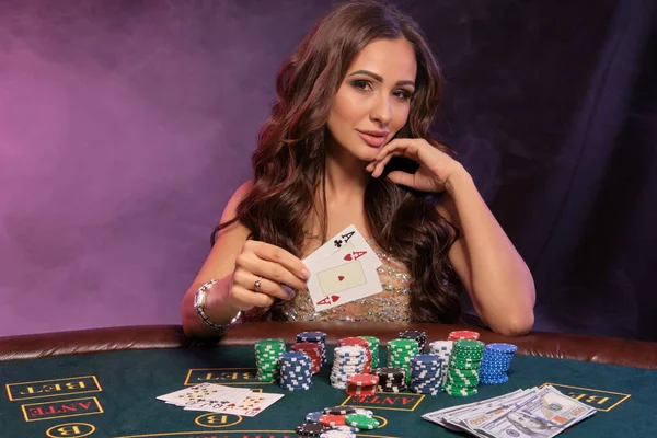 Flicka spelar poker, kasino. Visar kort, sitter vid bordet med högar av marker och pengar. Svart, rök bakgrund, färgglada bakljus. Närbild. — Stockfoto