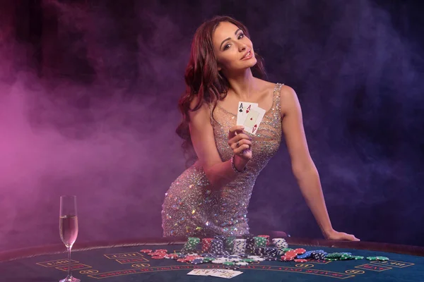 Menina jogando poker, cassino. Mostrando cartões, posando à mesa com pilhas de fichas e dinheiro. Preto, fundo de fumaça, backlights coloridos. Close-up. — Fotografia de Stock