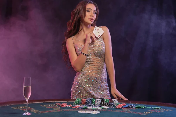 Gadis bermain poker, kasino. Menampilkan kartu, berpose di meja dengan tumpukan chip dan uang. Hitam, latar belakang asap, lampu belakang berwarna-warni. Close-up. — Stok Foto