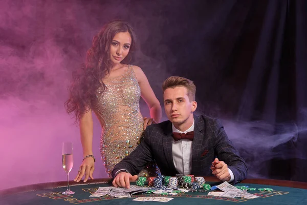 Ο τύπος παίζει πόκερ στο καζίνο, στο τραπέζι με στοίβες από μάρκες και κάρτες. Γιορτάζει τη νίκη με γυναίκα. Μαύρο, φόντο καπνού. Κοντινό πλάνο. — Φωτογραφία Αρχείου