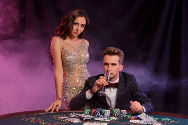 Guy está jugando al póquer en el casino, en la mesa con montones de fichas y cartas en él. Celebra ganar con la mujer. Fondos negros de humo. Cierre. — Foto de Stock