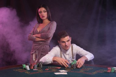 Kumarhanede poker oynayan bir adam, üzerinde bir yığın fiş ve kartla masada oturuyor. Kadınlarla kazanmayı kutluyoruz. Siyah, dumanlı arka plan. Yakın plan..