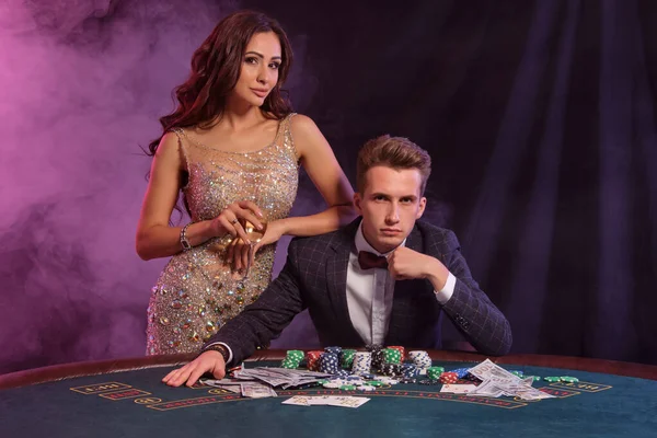 Guy is playing poker at casino, at table with stacks of chips and cards on it. He celebrating win with female. Black, smoke background. Close-up. — Stock Photo, Image