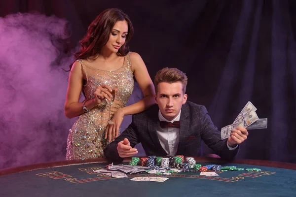 Guy holding cash, play poker at casino, sitting at table with stacks of chips, cards. Celebrating win with female. Black, smoke background. Close-up. — Stock Photo, Image