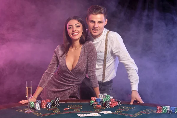 Guy in white shirt está jugando al póquer en el casino, en la mesa con montones de fichas en él. Celebra ganar con la mujer. Fondos negros de humo. Cierre. — Foto de Stock