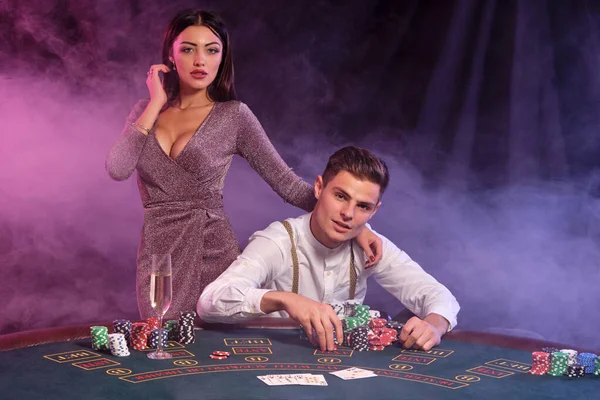 Hombre jugando al póquer en el casino, sentado en la mesa con montones de fichas y cartas en él. Celebrar la victoria con la mujer. Fondos negros de humo. Cierre. — Foto de Stock