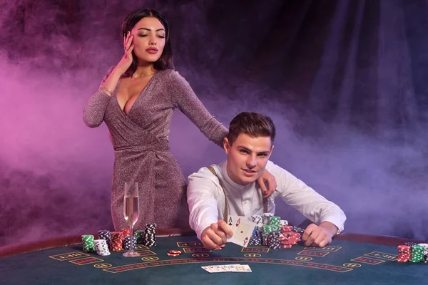 El hombre sostiene cartas, jugando al póquer en el casino, sentado en la mesa con montones de fichas en él. Celebra ganar con la mujer. Fondos negros de humo. Cierre. — Foto de Stock