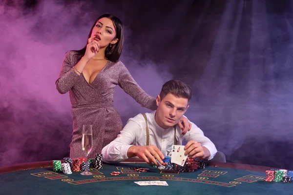 El hombre sostiene cartas, jugando al póquer en el casino, sentado en la mesa con montones de fichas en él. Celebra ganar con la mujer. Fondos negros de humo. Cierre. — Foto de Stock